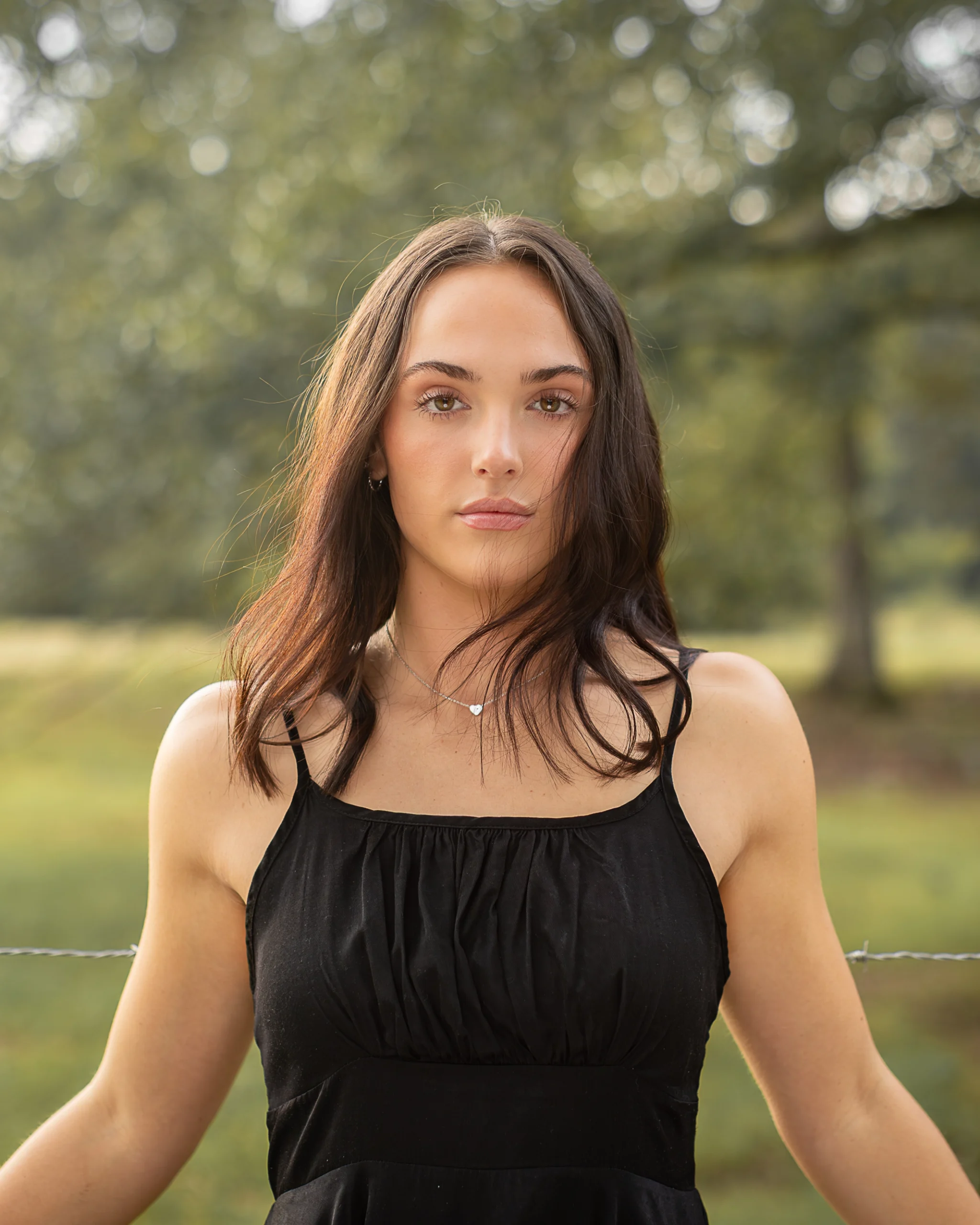 1 A woman with long brown hair wearing a black sleeveless dress stands outdoors in front of a blurred, green background.