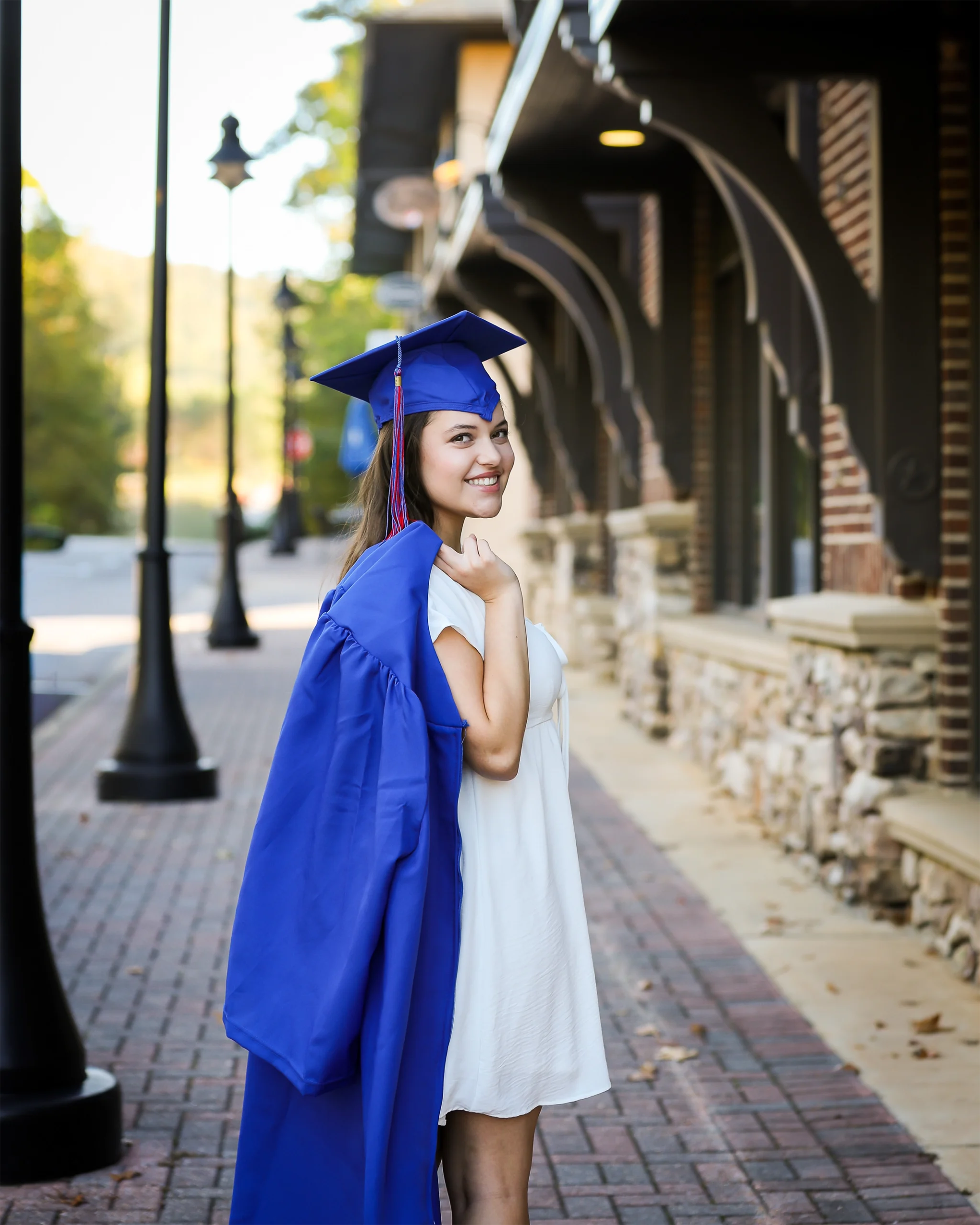 2 A young woman in a white dress stands on a brick path, smiling and holding her blue graduation gown. Street lamps and trees line the background. Celebratory mood.