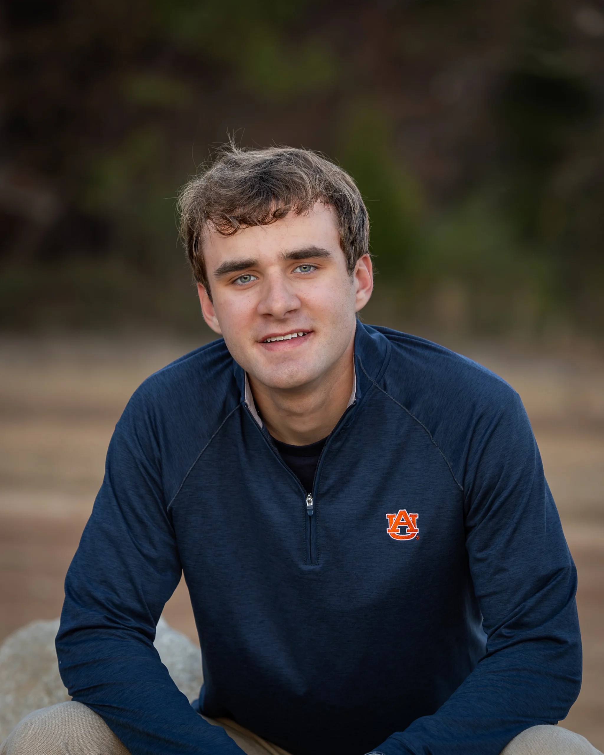3 Young man in a navy Auburn University quarter-zip sweater smiles gently, seated outdoors. The background is a blurred natural setting, conveying a calm tone.