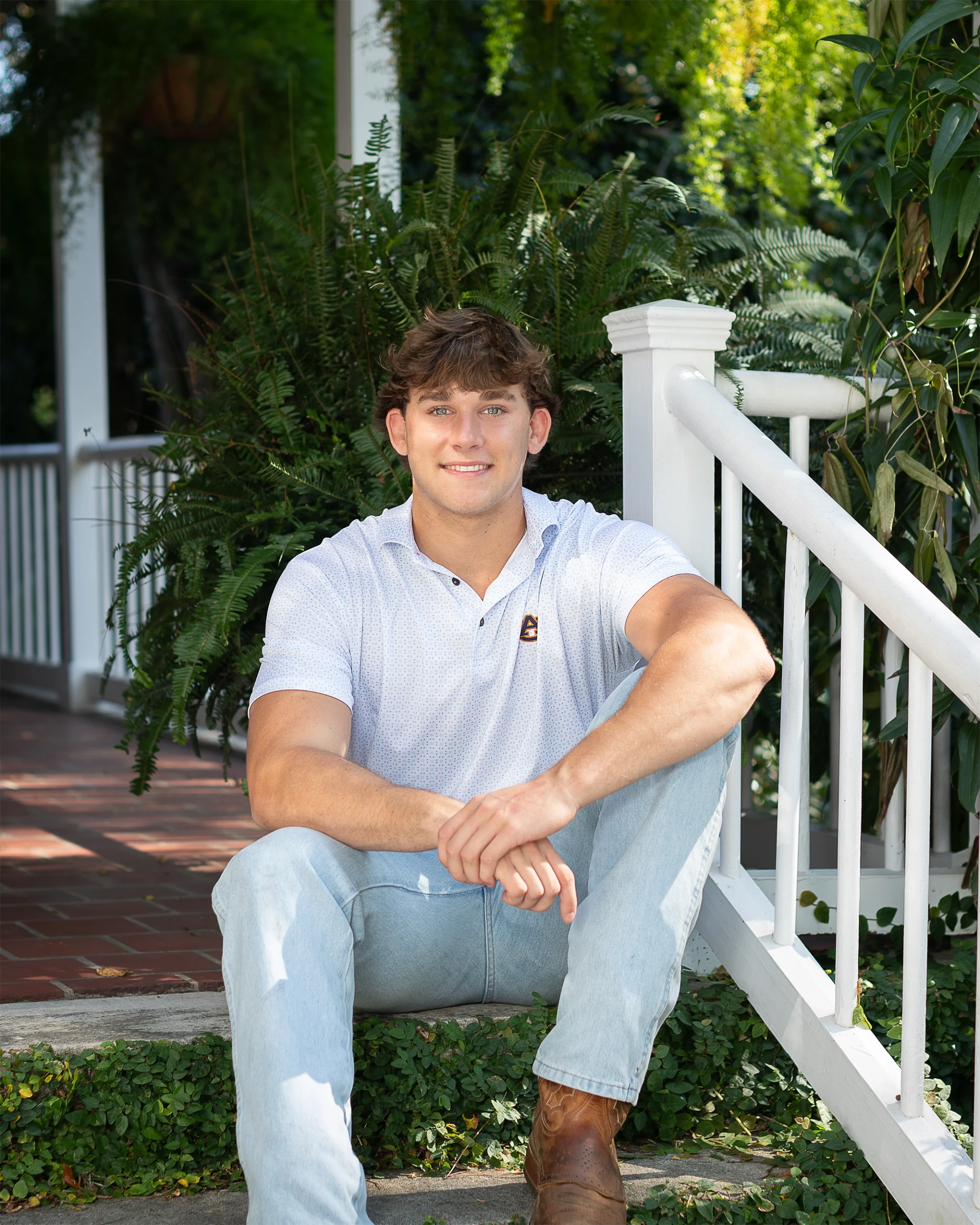 4 Young man sitting on porch steps with greenery in the background. He wears a white shirt, light jeans, and brown boots, smiling warmly at the camera.