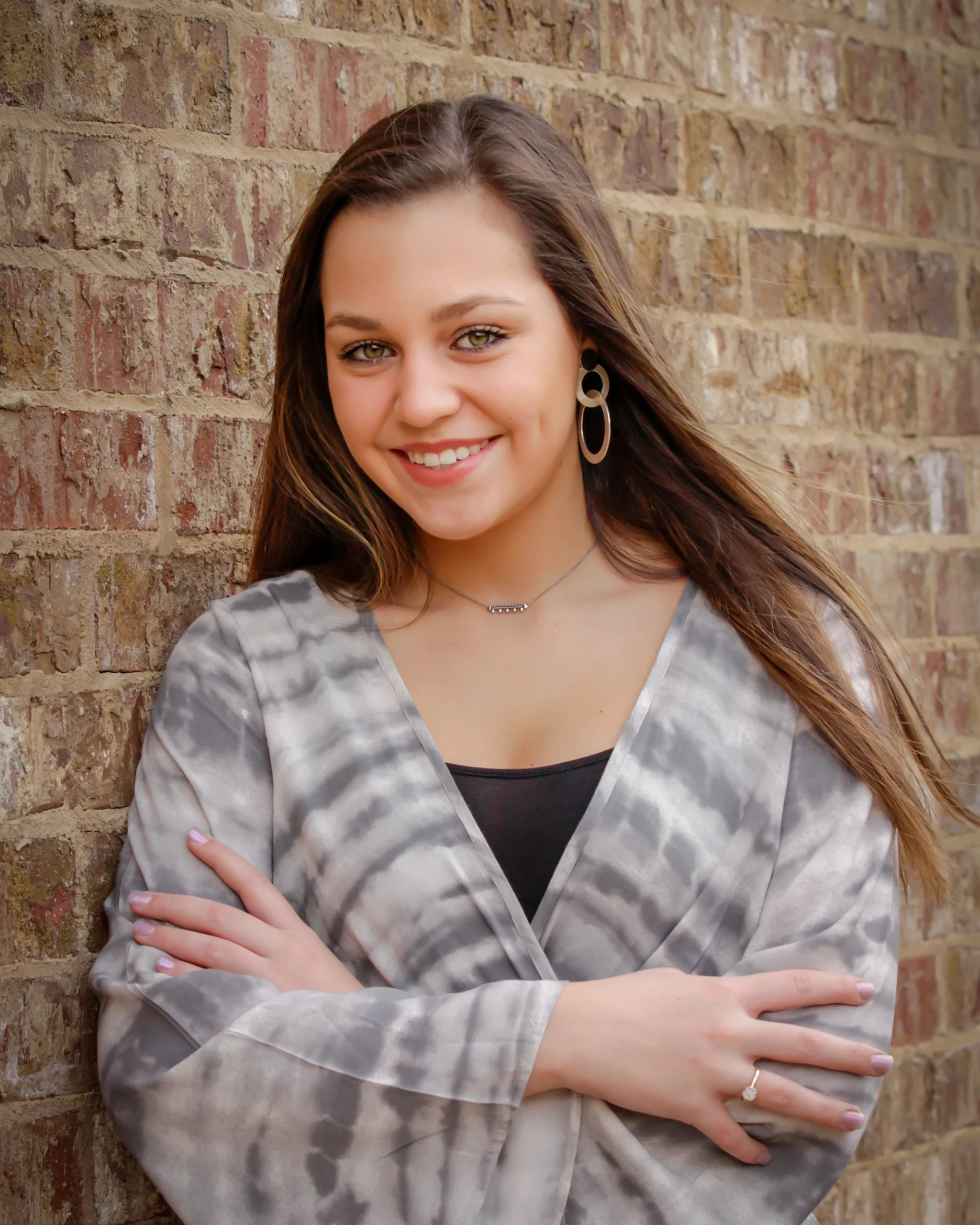 5 Young woman smiling confidently with crossed arms, wearing a gray tie-dye shirt and large hoop earrings, leaning against a brick wall.