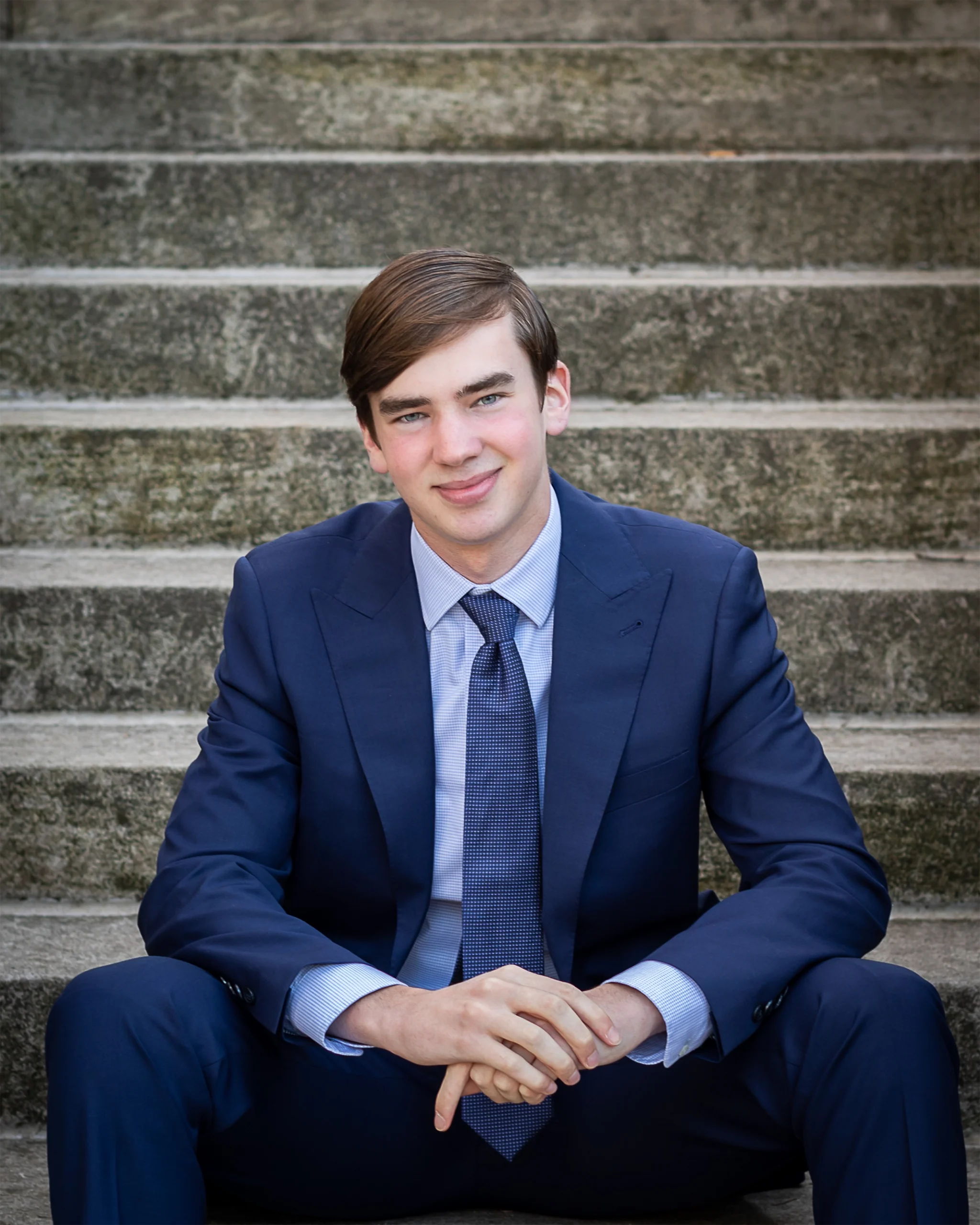 6 Young man in a blue suit sits confidently on stone steps, smiling. The setting feels professional and calm, conveying a sense of ambition and poise.